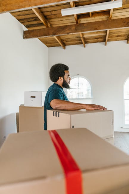 A man with dark skin, curly black hair, and a beard is seen in an unfinished interior space with a wooden ceiling and large arched windows, standing beside several closed cardboard boxes of varying sizes, some with red packing straps. He is wearing a blue T-shirt and appears to be engaged in a home relocation process, possibly preparing to load the boxes onto a vehicle or move them within the property. The boxes are stacked and arranged on the floor or a flat surface, indicative of packing or unpacking activities associated with furniture transport and moving services. The room has natural lighting coming through the windows, and the overall setting depicts an early stage of packing or unpacking during a house move, with the focus on efficient handling and organization of belongings, as handled by professional removals companies like Man with Van Kingston Vale.