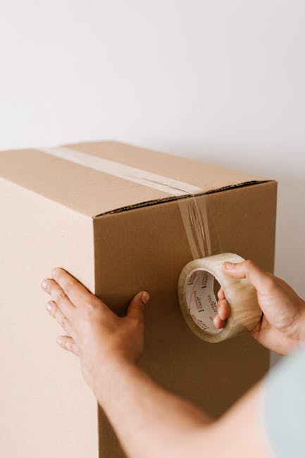 A person’s hands are visible, with one hand stabilising a large cardboard box and the other hand holding packing tape, preparing to seal the box. The cardboard box is plain, light brown, and appears to be filled with household items for moving. The tape is clear and being applied along the top seam of the box to secure it for transportation. The background is a plain, neutral-colored wall, indicating an indoor setting likely within a home during packing for a relocation. The scene illustrates the packing and moving process, potentially carried out by a professional service like Man with Van Kingston Vale, as part of home relocation or furniture transport. The image captures the careful preparation involved in house removals, emphasizing the importance of secure packing materials such as boxes and tape in ensuring safe transport of belongings.