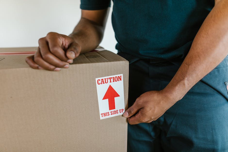 A man with dark skin, curly black hair, and a beard is seen in an unfinished interior space with a wooden ceiling and large arched windows, standing beside several closed cardboard boxes of varying sizes, some with red packing straps. He is wearing a blue T-shirt and appears to be engaged in a home relocation process, possibly preparing to load the boxes onto a vehicle or move them within the property. The boxes are stacked and arranged on the floor or a flat surface, indicative of packing or unpacking activities associated with furniture transport and moving services. The room has natural lighting coming through the windows, and the overall setting depicts an early stage of packing or unpacking during a house move, with the focus on efficient handling and organization of belongings, as handled by professional removals companies like Man with Van Kingston Vale.