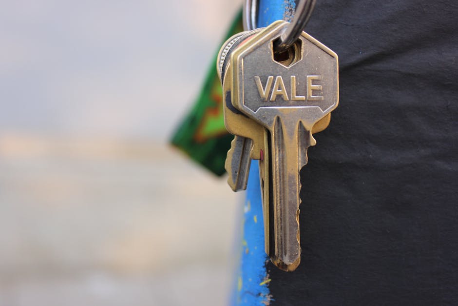 A close-up image showing a set of metallic keys hanging from a keyring, with one key prominently engraved with the name 'VALE'. The keys are attached to a person wearing a dark-colored jacket or coat,