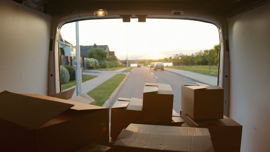 View from inside a moving van during a home relocation, with several cardboard boxes of varying sizes stacked at the back, some with flaps open. The boxes are packed with packing materials like paper or bubble wrap. The van's open rear door reveals a suburban street scene outside, featuring a paved road with parked cars, houses with gardens, street lamps, and greenery under warm evening sunlight. Inside the van, there is subdued lighting, highlighting the boxes ready for transport or unloading. This scene captures the loading process typical of furniture transport and packing during a house removal, illustrating professional logistics managed by Man with Van Kingston Vale, with the focus on organized packing for a smooth move in Kingston Vale.