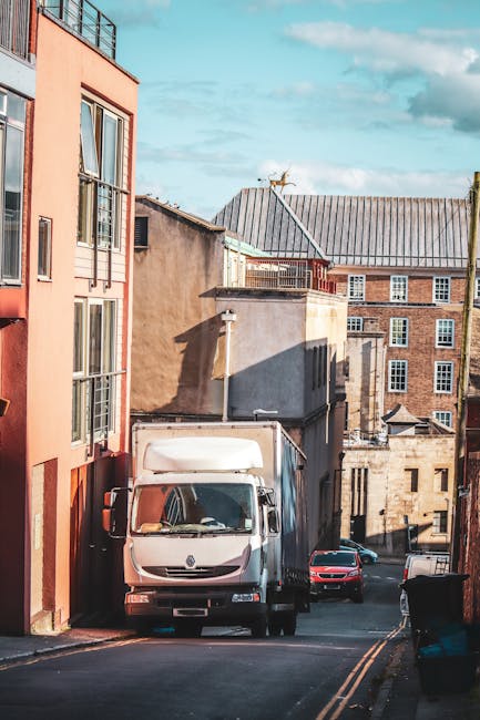 A white moving van with a closed cargo area is parked on a narrow, paved street in Kingston Vale, surrounded by residential buildings of various architectural styles and materials, including brick and stucco. The van is positioned alongside a multi-storey apartment building with large windows and balconies. On the street, a few vehicles, including a red car, are parked nearby. The scene is illuminated by natural daylight, casting shadows on the buildings and street, which suggests active home relocation or furniture transport preparations involving [COMPANY_NAME], as part of the house removals and packing process. Street-level details such as a waste bin and the pavement markings are also visible, reflecting typical urban moving logistics environment.