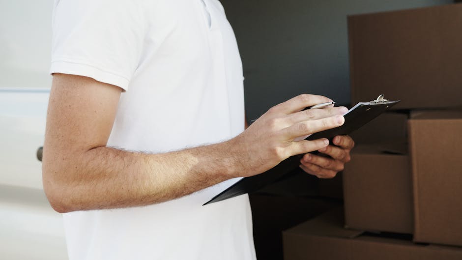Two moving staff members, wearing black T-shirts and caps with the logo 'RAC' and the wording 'High Quality Moving Service', are outside a modern apartment building during a house removal. They are lo