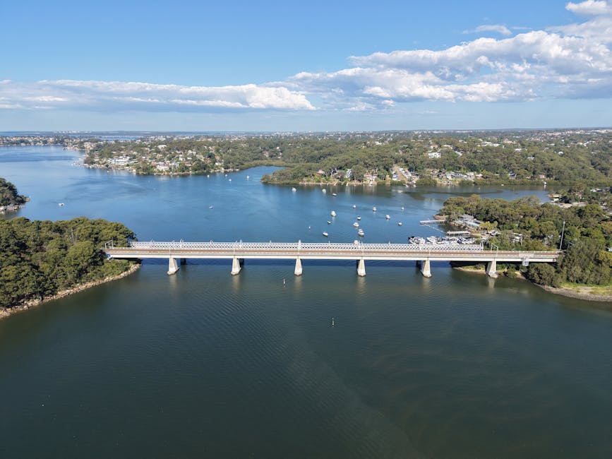 An aerial view of Kingston Bridge spanning across a wide river, with several small boats anchored on the water and lush greenery on either side of the riverbanks. In the background, there are urban areas with numerous residential buildings and trees, under a partly cloudy sky with sunlight illuminating the scene. The bridge appears to be a concrete structure with supporting columns in the water, and a parked vehicle is visible on the bridge's road surface. This image exemplifies the scenic environment surrounding Kingston Vale, where home relocation and furniture transport services by Man with Van Kingston Vale are often coordinated for smooth moving logistics across the area.