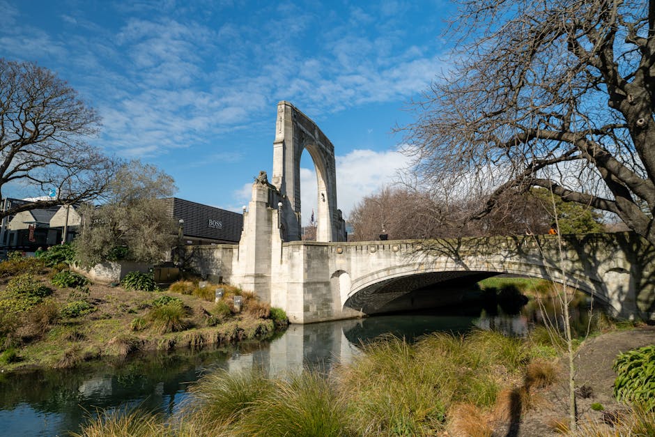 A historic stone bridge with an arched design and a large, open central arch spans a calm river in an outdoor setting. The bridge features a tall, decorative arch rising above the main structure, supported by stone piers and parapets. Surrounding the bridge are leafless trees and landscaped greenery, including bushes and grasses, indicating a season with sparse foliage. The sky is partly cloudy with patches of blue, providing natural daylight that highlights the weathered stone surface of the bridge. In the background, there are modern commercial buildings and a visible black van parked along the roadside. The scene captures the tranquil environment near Kingston Vale, with the bridge situated adjacent to the water's edge, where the river flows beneath the protected arch. This setting could be relevant for visualizing the transportation or access points involved in house removals and packing for a home relocation.