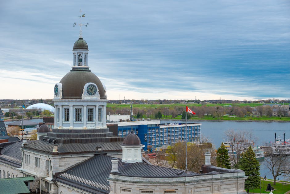 An aerial view of Kingston Bridge spanning across a wide river, with several small boats anchored on the water and lush greenery on either side of the riverbanks. In the background, there are urban areas with numerous residential buildings and trees, under a partly cloudy sky with sunlight illuminating the scene. The bridge appears to be a concrete structure with supporting columns in the water, and a parked vehicle is visible on the bridge's road surface. This image exemplifies the scenic environment surrounding Kingston Vale, where home relocation and furniture transport services by Man with Van Kingston Vale are often coordinated for smooth moving logistics across the area.