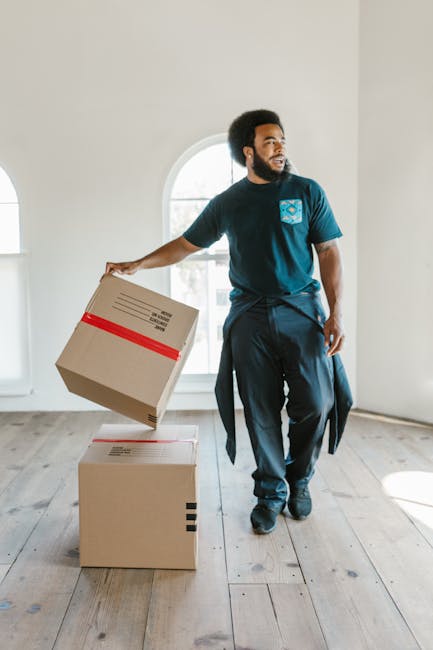 A man with dark curly hair and a beard, wearing a navy blue uniform with a logo on the chest, is inside a bright, spacious room with white walls and a large arched window. He is holding a medium-sized cardboard box with red tape around it, preparing for a home relocation. The room has a light wooden floor and minimal furnishings, with another sealed cardboard box placed on the floor nearby. Natural light floods the space through the window, illuminating the scene as part of a furniture transport or packing process. This image exemplifies professional removal services, such as those offered by Man with Van Kingston Vale, during a house move, with emphasis on careful handling and structured packing during the loading process involved in house removals in Kingston Vale. The visual highlights the careful handling of packaging materials and the systematic approach to furniture transport within a residential setting.