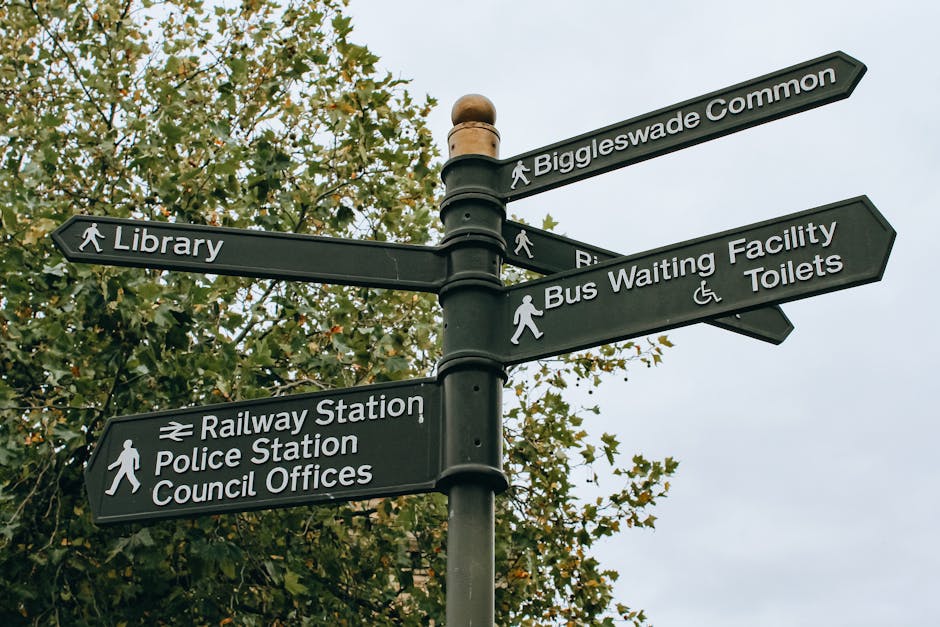 A black multi-directional street sign post with six signs attached, each pointing to different local landmarks and facilities including Biggleswade Common, the Library, Railway Station, Police Station, Council Offices, and Bus Waiting Facility and Toilets, situated outdoors against a backdrop of green trees and a cloudy sky. The signs feature white lettering and icons representing pedestrians and accessibility, indicating nearby points of interest within the town. This signage is part of a typical urban environment used for navigating residential and public service areas in Kingston Vale. The context suggests that [COMPANY_NAME], a professional removals service, is familiar with moving logistics in such local settings, highlighting the importance of clear signage during the moving process and home relocation activities.