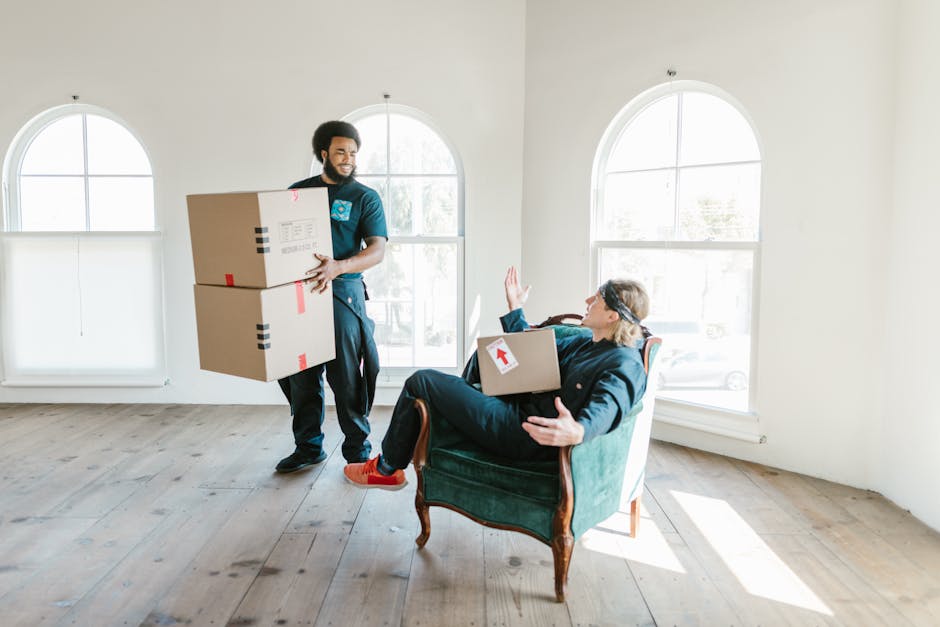 A professional moving assistant from Man with Van Kingston Vale is inside a bright, minimally furnished room with large arched windows and natural light. The assistant, dressed in dark clothing, is st