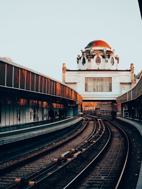 A photograph of a train station platform with curved metal railings and a set of railway tracks leading towards a historic white building with a domed roof and decorative architectural details in the background. The platform edge is lined with glass panels and safety barriers, and there are various cardboard boxes, moving blankets, and plastic wrapping materials placed on the platform, indicating a home relocation process. The scene is illuminated by natural daylight, with the sky visible above the building. The image captures the logistical aspect of furniture transport and packing within a house removal context, aligning with services offered by Man with Van Kingston Vale during local moves such as the Norbiton station flat removals success story in Kingston Vale.