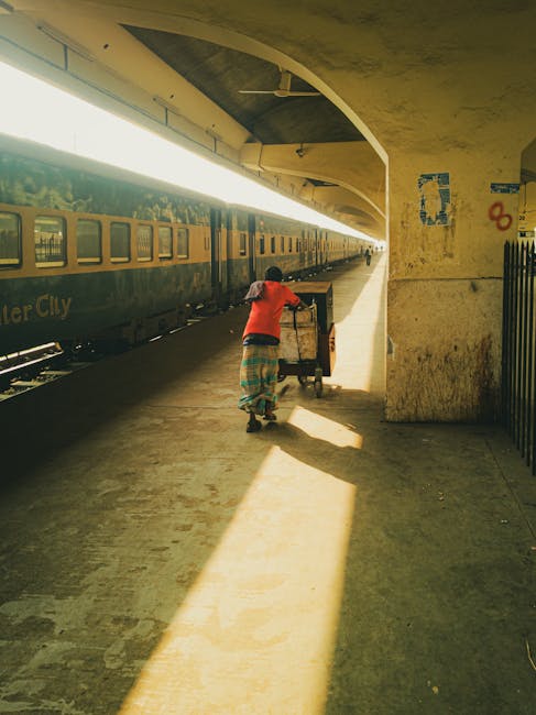 A person wearing a red shirt and colorful, patterned clothing pushes a wheeled trolley along the platform of Norbiton station, Kingston Vale, during daylight hours. The platform is partially shaded by an arched concrete overhead structure, with sunlight streaming through the open side, casting long shadows on the paved surface. A green train with closed windows is parked on the adjacent track, indicating an ongoing home relocation process involving furniture transport and packing. To the right, there is a black metal fence, and behind the person, a large concrete pillar supports the station structure. The scene captures the movement of belongings during a house removal, with visible elements of packing and loading in a typical railway station environment, reflecting the logistical aspects of relocations managed by professional removals services like Man with Van Kingston Vale.
