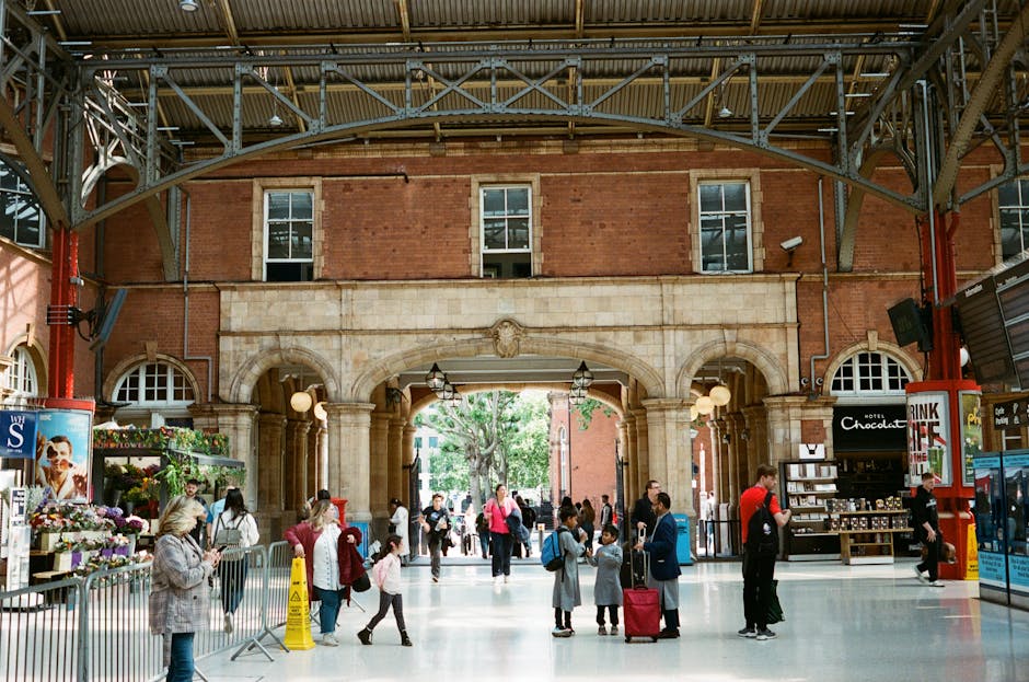A photograph of a train station platform with curved metal railings and a set of railway tracks leading towards a historic white building with a domed roof and decorative architectural details in the background. The platform edge is lined with glass panels and safety barriers, and there are various cardboard boxes, moving blankets, and plastic wrapping materials placed on the platform, indicating a home relocation process. The scene is illuminated by natural daylight, with the sky visible above the building. The image captures the logistical aspect of furniture transport and packing within a house removal context, aligning with services offered by Man with Van Kingston Vale during local moves such as the Norbiton station flat removals success story in Kingston Vale.