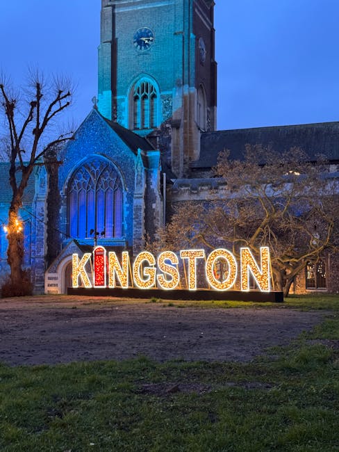 A large illuminated sign spelling 'KINGSTON' in bright, multicoloured light bulbs is positioned on the grass in front of a historic stone church with pointed Gothic windows. The church is lit with blue lighting, highlighting its tall tower and arched windows, and is surrounded by leafless trees. The scene appears to be taken at dusk or night, with a clear sky overhead. This setting suggests a location in Kingston, possibly during a local event or holiday, with ongoing hotel or house removal preparations by Man with Van Kingston Vale, as part of their house removals and relocation services in the area.