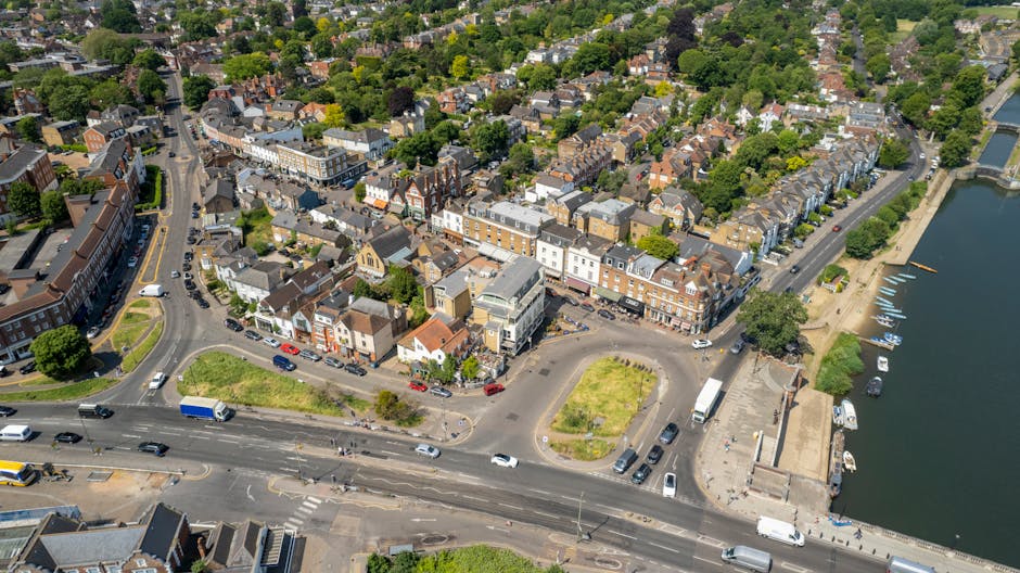 An aerial view of a residential area near a river showing a mix of terraced and detached houses with chimneys, surrounded by trees and greenery. A road with multiple lanes runs along the front of the properties, with several vehicles including cars, vans, and a small truck moving along it. Near the road, there are parking areas with additional parked cars and some open spaces with grass patches and small trees. The image captures a scene of home relocation activity, with a focus on the urban environment typical of Kingston Vale, while emphasizing the context of house removals and furniture transport services offered by Man with Van Kingston Vale. The river on the right side of the image has boats docked along the quay, and the overall setting includes natural and built scenery, with good daylight illuminating the entire area.