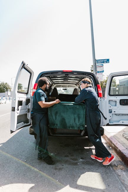 Two movers from Man with Van Kingston Vale are engaged in a furniture transport operation during a home relocation in the Richmond Park area. They are standing at the rear of a black delivery van with its doors open, carefully lifting and guiding a large, dark green upholstered piece of furniture into the vehicle. The interior of the van contains additional packing material and possibly other household items prepared for safe moving. The movers are dressed in dark uniforms, one wearing glasses and sneakers, and both are focused on the loading process. The scene takes place outdoors on a paved driveway, with a clear sky overhead and a street sign visible in the background. Nearby, a segment of the pavement and curb can be seen, along with a roadside area typical of Kingston Vale. The image captures a key moment in a professional furniture transport and packing service, illustrating careful handling and organized loading as part of a comprehensive removals service.