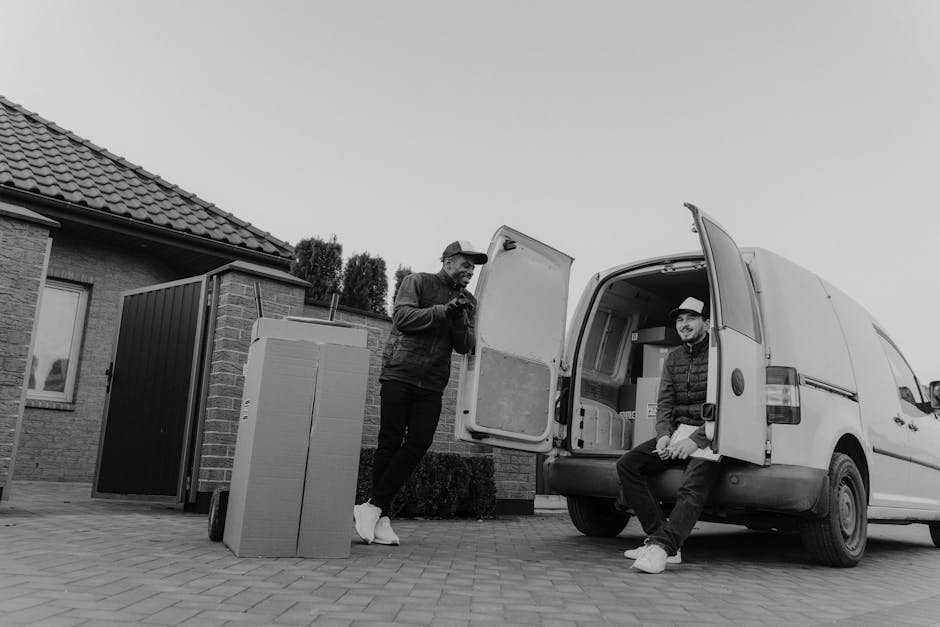 Two men engaged in a furniture transport process outside a residential property, with one standing next to a white delivery van and the other sitting on the van’s open rear door. The standing man, wearing a cap, jacket, and sneakers, appears to be inspecting or communicating, while the seated man, also in casual attire and a hat, is resting with his hands on his lap. In the foreground, a large cardboard box and a tall, narrow cardboard container are placed on the paved driveway, ready for loading. The background reveals a brick house with a tiled roof, a small garden area, and a black fencing panel. The scene is set during daylight hours with even lighting, capturing the typical elements of a house removal service focused on furniture transport and packing logistics, as executed by Man with Van Kingston Vale, in the context of a home relocation in the Richmond Park area.