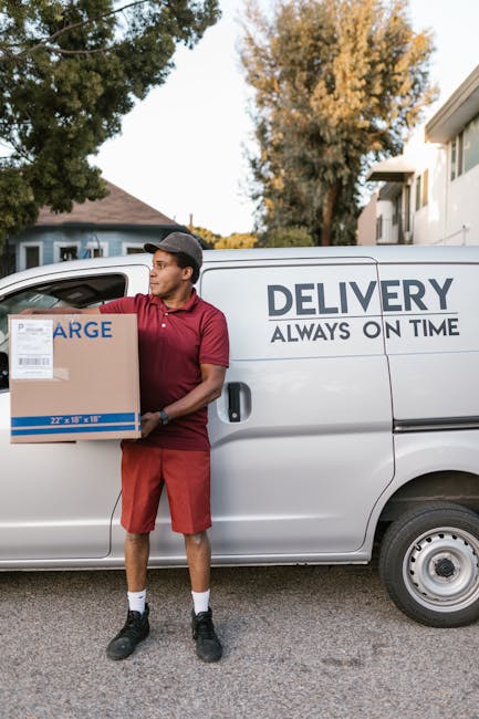 Two movers from Man with Van Kingston Vale are engaged in a furniture transport operation during a home relocation in the Richmond Park area. They are standing at the rear of a black delivery van with its doors open, carefully lifting and guiding a large, dark green upholstered piece of furniture into the vehicle. The interior of the van contains additional packing material and possibly other household items prepared for safe moving. The movers are dressed in dark uniforms, one wearing glasses and sneakers, and both are focused on the loading process. The scene takes place outdoors on a paved driveway, with a clear sky overhead and a street sign visible in the background. Nearby, a segment of the pavement and curb can be seen, along with a roadside area typical of Kingston Vale. The image captures a key moment in a professional furniture transport and packing service, illustrating careful handling and organized loading as part of a comprehensive removals service.