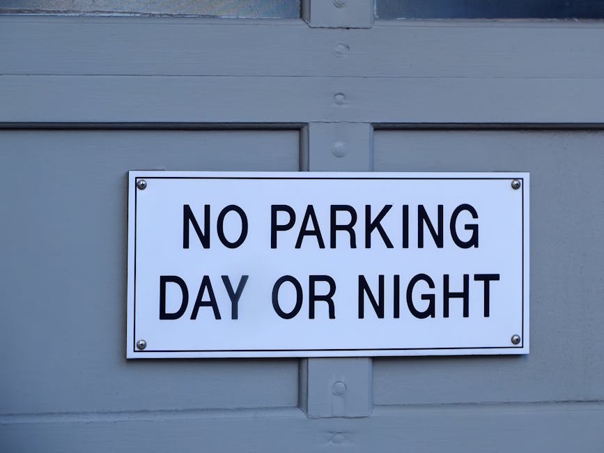 A rectangular white metal sign with black uppercase text reading 'NO PARKING DAY OR NIGHT' attached to a grey wooden building with visible horizontal panels and a central vertical support. The sign is fastened with four screws, one in each corner. The background shows part of a doorway or window frame below the sign, and the lighting is neutral, indicating daytime. The image pertains to parking restrictions relevant to house removals and local parking rules, as might be considered by companies like Man with Van Kingston Vale during home relocation or furniture transport in Kingston Vale, close to the entrance of a property.