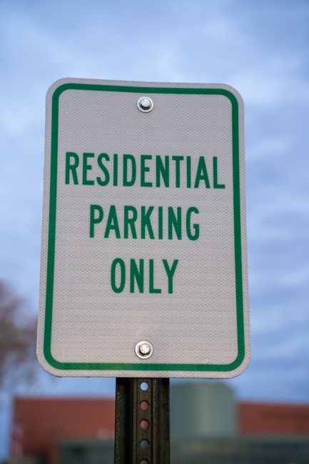 A close-up of a rectangular green and white sign mounted on a metal pole, indicating 'RESIDENTIAL PARKING ONLY' in bold green capital letters. The sign is outside a property, with a clear blue sky and some blurred building rooftops visible in the background. This type of signage is relevant for home relocations and furniture transport, as it highlights parking restrictions that removals companies like Man with Van Kingston Vale need to consider when planning the loading process and vehicle access during house removals in Kingston Vale.